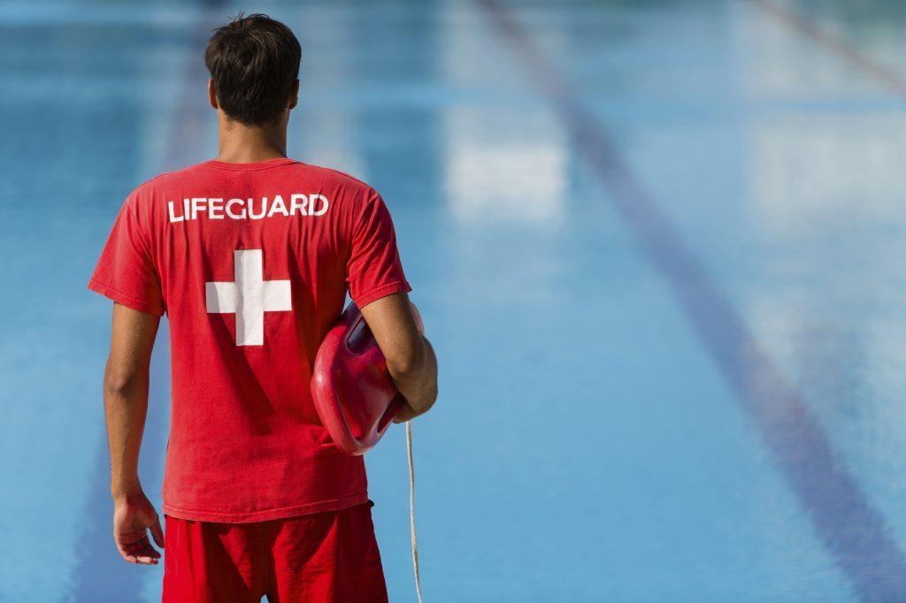 Rear view of male lifeguard with emergency equipment in red uniform watching swimming pool.